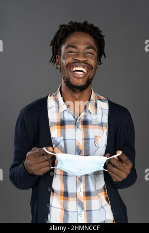 Homme afro-américain tenant un masque médical dans les mains souriant dans l'appareil photo. Un jeune homme heureux ou fatigué qui prend un masque médical. Homme africain avec un masque médical entre les mains. Isolé sur gris. Banque D'Images