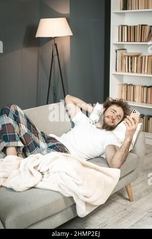 Malade jeune homme en pyjama dormant sur le canapé regardant un smartphone ou ayant un appel vidéo . Beau jeune homme barbu dormant sur le canapé après de longues heures de travail tard le soir. Banque D'Images