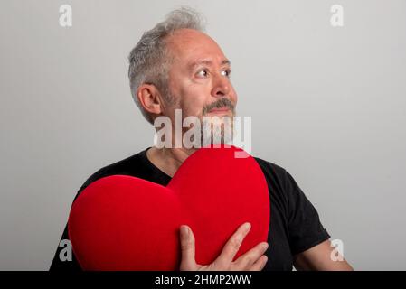 Beau homme mi-âge gris-poil et barbu tenant un coeur rouge, isolé fond gris. Concept de la Saint-Valentin Banque D'Images