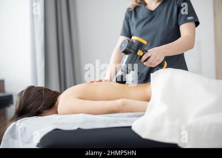 La femme fait un massage du dos du patient à l'aide d'un masseur à percussion Banque D'Images