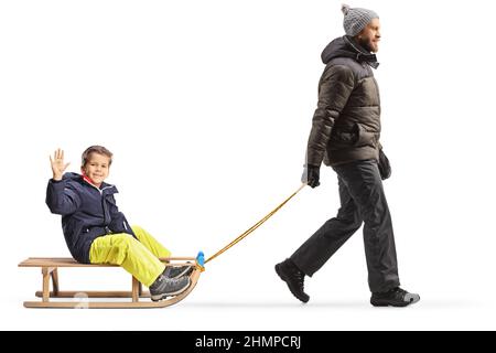 Photo pleine longueur d'un père en vêtements d'hiver marchant et tirant un garçon sur un traîneau isolé sur fond blanc Banque D'Images