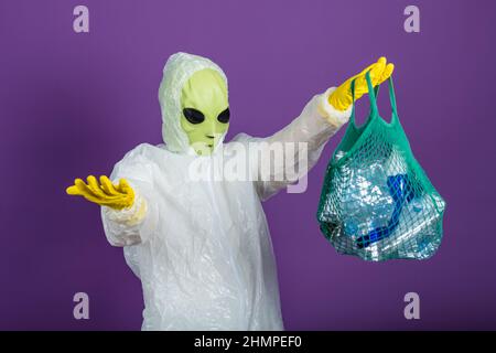 Drôle d'homme dans un masque étranger dans un costume blanc tient un sac en maille avec des bouteilles en plastique et répand ses mains sur un fond violet, concept de tri des déchets Banque D'Images