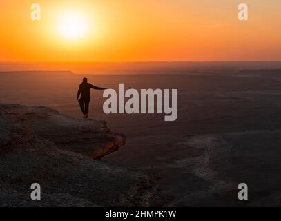 Homme avec une caméra sur un bâton de selfie marchant le long d'une falaise au coucher du soleil, le bord du monde (Jebel Fihrayn), Arabie Saoudite Banque D'Images