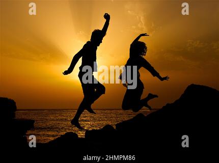 Silhouette d'un homme et d'une femme sur la plage sautant dans l'air au coucher du soleil, Costa Teguise, Lanzarote, îles Canaries, Espagne Banque D'Images