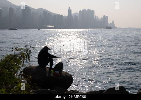 Silhouette d'homme assis sur le rocher, pêche au coucher du soleil au port de Victoria avec paysage urbain de Hong Kong derrière Banque D'Images