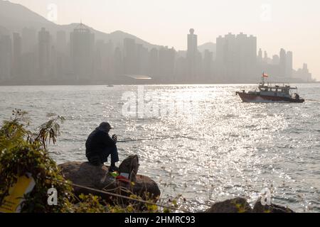 Silhouette d'homme assis sur le rocher, pêche au coucher du soleil au port de Victoria avec paysage urbain de Hong Kong derrière Banque D'Images