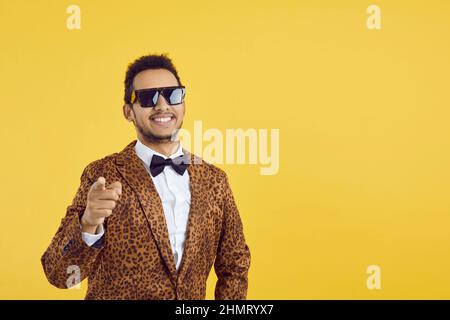 Joyeux gars noir dans des verres frais et costume funky pointe le doigt sur l'appareil photo sur l'arrière-plan de l'espace de copie Banque D'Images