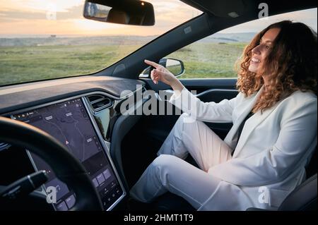 POV jeune femme d'affaires assis dans le siège passager à côté du conducteur et pointant le doigt devant elle. Arrière-plan des champs et du coucher du soleil. Banque D'Images