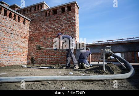 Deux travailleurs en cours d'application du mélange de ciment fourni par une pompe à mortier mobile sur la surface de travail. Installation d'une table à plancher semi-sec à l'extérieur sous ciel ouvert bleu. Banque D'Images