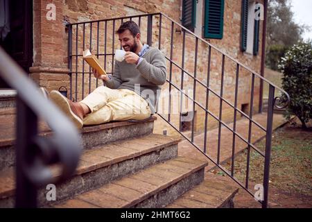Un jeune homme est en train de lire un livre intéressant tout en étant assis sur le porche et en appréciant un thé chaud sur une belle journée. Ville, extérieur, repos Banque D'Images