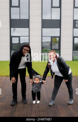 Jeunes parents multiethniques marchant dans le parc avec un petit fils, passant des week-ends à l'extérieur. Mère élégante et père charmant aidant un enfant mignon avec des firs Banque D'Images