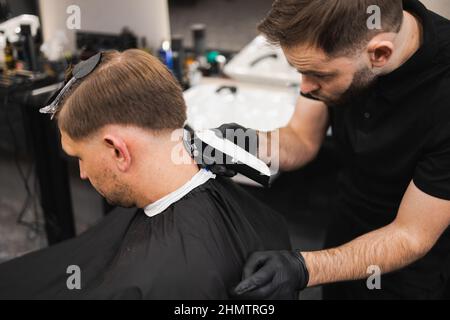 Jeune homme dans un salon de coiffure. Le coiffeur coupe les cheveux depuis l'arrière de la tête du client à l'aide d'une tondeuse électrique Banque D'Images