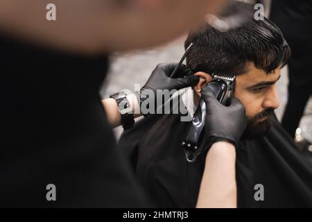 Gros plan des mains d'un coiffeur en gants de latex noir coupant les cheveux sur la tête d'un homme avec une tondeuse Banque D'Images