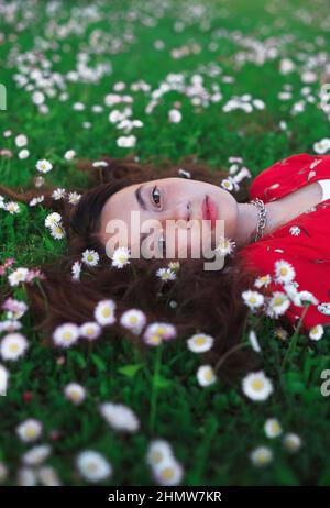 Jeune belle fille en robe rouge repose sur l'herbe de printemps fraîche avec de petites fleurs blanches. Banque D'Images