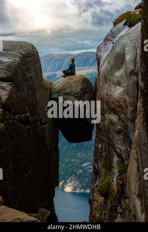 La femme est assise dans une posture méditative au sommet de Kjeragbolten - bloc coincé dans la crevasse de la montagne à 984 mètres au-dessus de la mer Banque D'Images