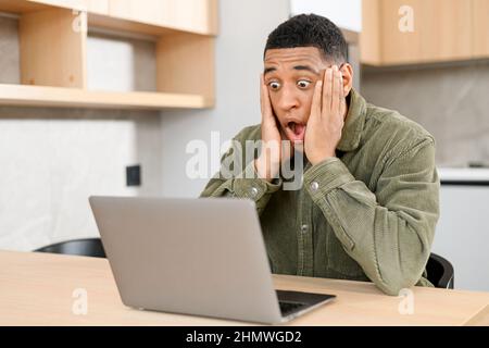 Excité surpris latin sombre-cheveux homme dans une chemise décontractée regardant à l'ordinateur portable. Choqué heureux entrepreneur multiracial masculin a reçu bonne affaire, e-mails avec offre d'emploi Banque D'Images