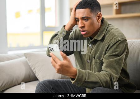 Un homme stressé en colère a une conversation d'appel vidéo désagréable, parlant sur le smartphone en ligne et arguant, tandis que s'assoit sur le canapé et regarde l'écran du téléphone avec une expression de visage frustrée Banque D'Images