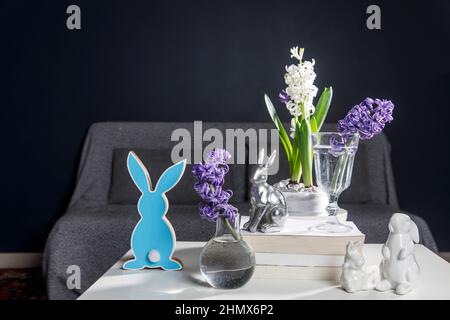 Figurines en céramique de lapins de Pâques de différentes tailles sur la table. Jacinthes bleu et blanc dans des tasses en verre sur fond sombre. Motif Pâques Banque D'Images