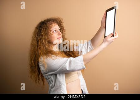 Photo de vue latérale d'une jeune femme à tête rouge tenant dans ses mains tablette et allant faire selfie sur fond d'or. Banque D'Images