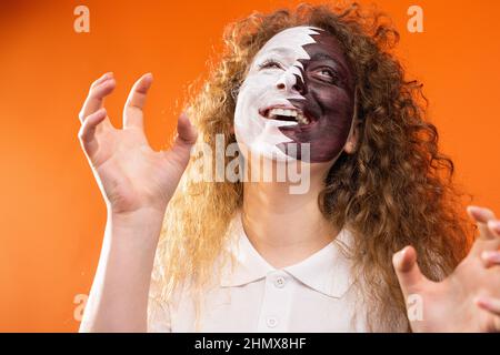 Jeune belle femme avec un visage heureux souriant en gardant les mains en position pour attraper une balle de football avec le visage peint dans le drapeau du Qatar. Banque D'Images