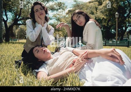 trois jeunes filles argentines douces et rétro, assises sur l'herbe, dans un parc public avec des arbres derrière elles, en regardant la caméra retr Banque D'Images