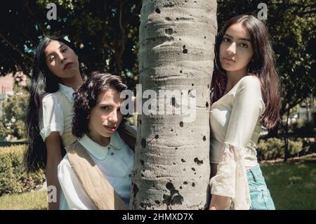 trois jeunes filles hispaniques argentines rétro de style vintage par une journée ensoleillée se posant autour d'un arbre dans un parc qui regarde la caméra Banque D'Images