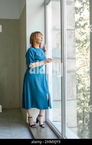 Belle femme mûre en robe bleue regardant à travers une fenêtre Banque D'Images