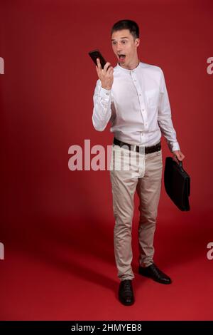 Un homme en colère dans une chemise blanche et un pantalon léger avec un sac à documents dans sa main hurle sur un téléphone portable sur le haut-parleur.Un homme stressé et déprimé sur fond rouge.Homme émotionnel Banque D'Images