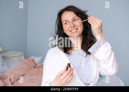 Concept beauté et soins de la peau. Le visage de la femme applique du sérum pour le visage de l'arbre à thé. Belle fille brune naturelle sans maquillage applique du sérum sur son visage avec une pipette. Dans un peignoir dans la chambre sur le lit Banque D'Images