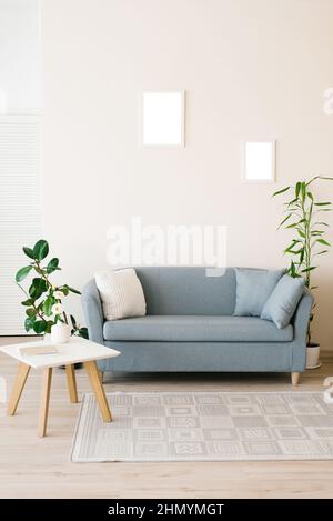 Un canapé bleu poussiéreux avec oreillers, une table basse blanche, des plantes en pot dans un salon lumineux et confortable. Cadres de maquette pour affiches sur le mur Banque D'Images
