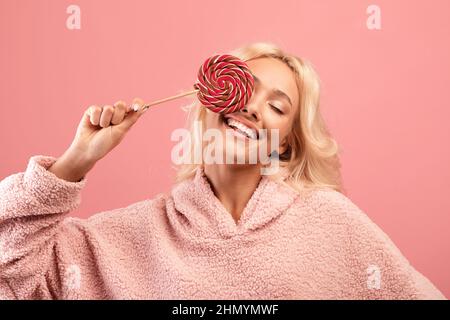 Jeune femme joyeuse tenant des bonbons sur le bâton, couvrant un oeil avec le lollipop et souriant, s'amusant sur fond rose Banque D'Images