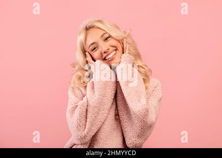 Portrait de jolie dame blonde touchant les joues dans l'excitation, debout heureux et souriant à la caméra, fond rose Banque D'Images