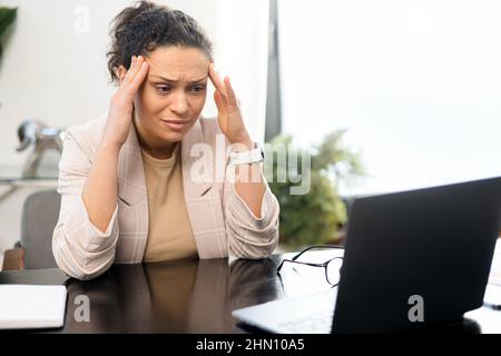 Inquiète femme d'âge moyen assis à la réception devant un ordinateur portable dans un bureau moderne, regarde l'écran et tient des temples, une employée de sexe féminin ressent un mal de tête, soucieuse de résoudre des tâches Banque D'Images