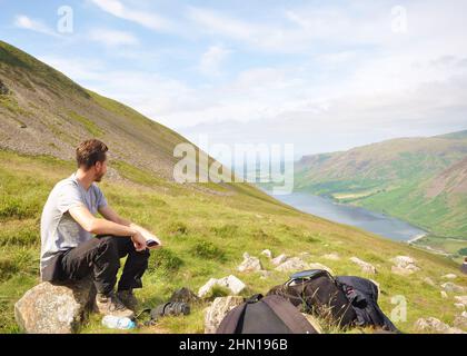 Randonneur en train de faire une pause tout en grimpant en montagne. Scafell Pike, Lake District, Royaume-Uni, vue vers Wast Water. Vue panoramique sur la montagne vers le lac avec de l'herbe Banque D'Images