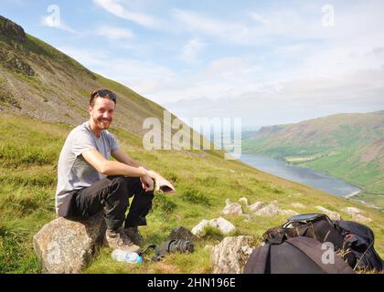 Randonneur en train de faire une pause tout en grimpant en montagne. Scafell Pike, Lake District, Royaume-Uni, vue vers Wast Water. Vue panoramique sur la montagne vers le lac avec de l'herbe Banque D'Images