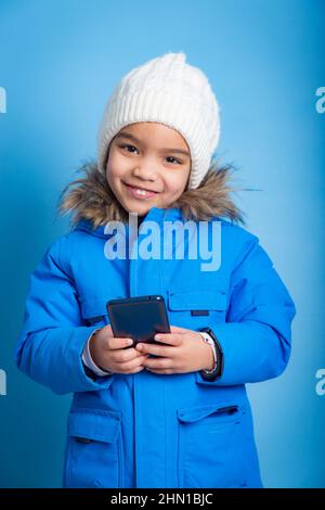 Petite fille tenant un smartphone portant un costume d'hiver sur fond bleu Banque D'Images