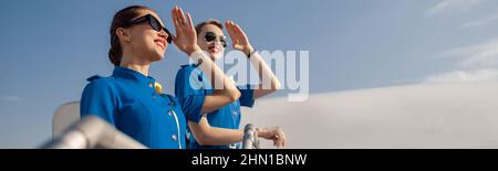 Portrait de deux élégantes hôtesses d'air en uniforme bleu et lunettes de soleil couvrant les yeux avec la main et regardant loin, debout ensemble sur un escalier d'air Banque D'Images