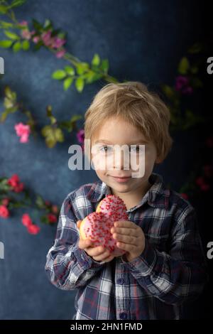 Mignon petit enfant blond, petit garçon d'âge préscolaire, mangeant le donut rose en forme de coeur, fait pour la Saint-Valentin, fond de Saint-Valentin Banque D'Images