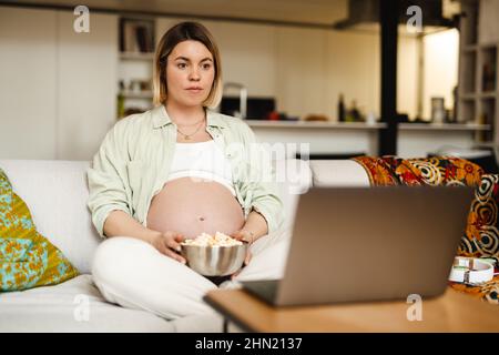 Une jeune femme enceinte mange du pop-corn et utilise un ordinateur portable tout en étant assise sur un canapé à la maison Banque D'Images