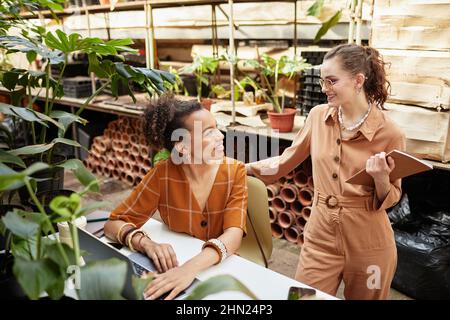 Deux jeunes jolies femmes consultant sur l'ordre dans la boutique en ligne tout en ayant la réunion par bureau à orangerie ou serre moderne Banque D'Images