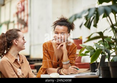 Jeune femme biraciale souriante gardenier et sa collègue caucasienne ayant discuté de la conception intérieure de leur nouvelle boutique de fleuristes Banque D'Images