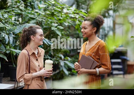 Bonne jeune femme avec des boissons consultant avec le fleuriste confiant ou concepteur de l'intérieur des magasins de fleurs tout en se tenant contre la plante verte Banque D'Images