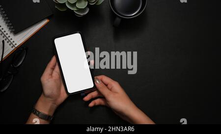 Prise de vue au-dessus de la tête, Une femme tient l'écran blanc du smartphone sur un arrière-plan noir moderne de l'espace de travail. Banque D'Images