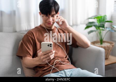 Concept technologique la personne qui porte des lunettes et des écouteurs assis sur le canapé gris et qui écoute de la musique sur son smartphone. Banque D'Images