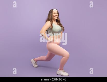 Gaie jeune femme européenne plus taille dans les vêtements de sport faire des fentes, des exercices de jambe isolés sur fond violet, profil Banque D'Images