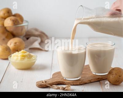 Lait de pomme de terre versé dans le verre sur fond de bois blanc. Verser le lait végétalien dans le verre, avec la purée de pommes de terre et les tubercules de pommes de terre sur le fond. Copier l'espace. Lait de pomme de terre fait maison à base de pommes de terre bouillies Banque D'Images