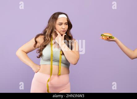 Femme pensive millénaire caucasienne de taille plus en vêtements de sport avec mètre ruban regarde à la main avec le hamburger Banque D'Images