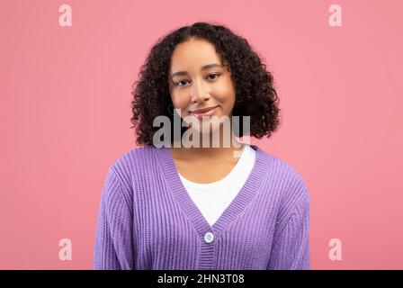 Jeune femme afro-américaine joyeuse avec un sourire léger et des cheveux bouclés regardant l'appareil photo sur fond rose studio Banque D'Images