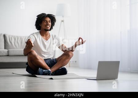Yoga domestique. Beau Black Guy méditant devant l'ordinateur portable à la maison Banque D'Images