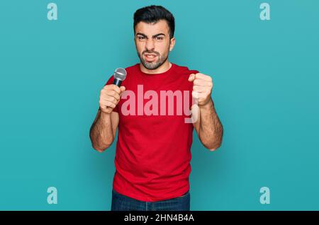 Homme hispanique avec chant la barbe chantant le chant en utilisant le microphone agacé et frustré criant avec la colère, hurlant fou avec la colère et la main levée Banque D'Images